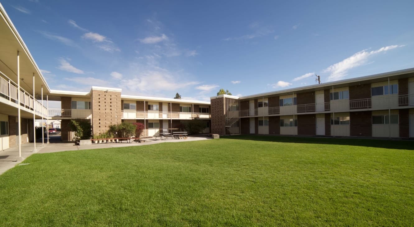 A large grassy courtyard at Cottonwood Rexburg is surrounded by two-story apartment buildings with balconies and exterior walkways under a blue sky with scattered clouds.