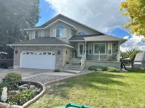 Two-story suburban house at 1555 S 630 E, Orem UT 84097, featuring a stone and siding exterior, attached two-car garage, front porch with white railing, landscaped yard with green grass, trees, and a partly cloudy sky in the background.