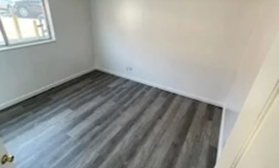 Freshly renovated and unfurnished, this small room at Parkway Apartments Orem features light gray walls, a window filling the space with natural light, and dark gray wood-style flooring.