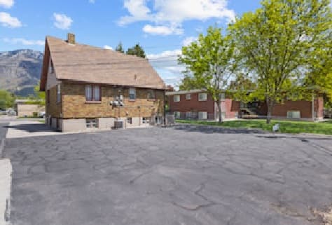 A small brick house with a steep roof sits beside an empty, cracked asphalt parking lot. Green trees and another red brick building are visible in the background, under a partly cloudy sky.