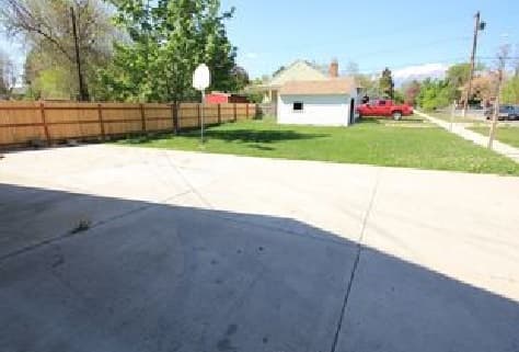 A concrete driveway at 591 E 100 N Provo features a basketball hoop on the side, beside a grassy yard, detached white garage, and red truck, all bordered by a wooden fence and trees.