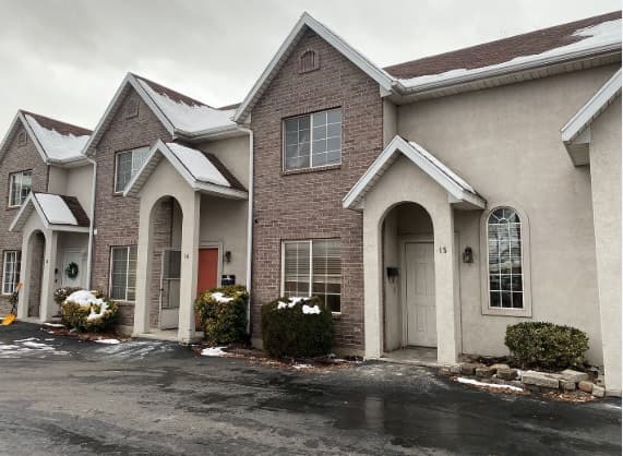 A row of two-story townhouses at 865 N 500 W #15 Provo features arched doorways and windows, some with brick accents. Bushes line the fronts, and traces of snow rest on the ground and rooftops beneath a cloudy sky.