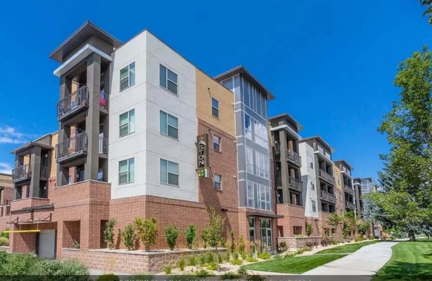 Modern four-story apartment building, The Aston at University Place Orem, features brick and light-colored siding, balconies, and large windows. A sidewalk, green grass, and trees are in the foreground under a clear blue sky.
