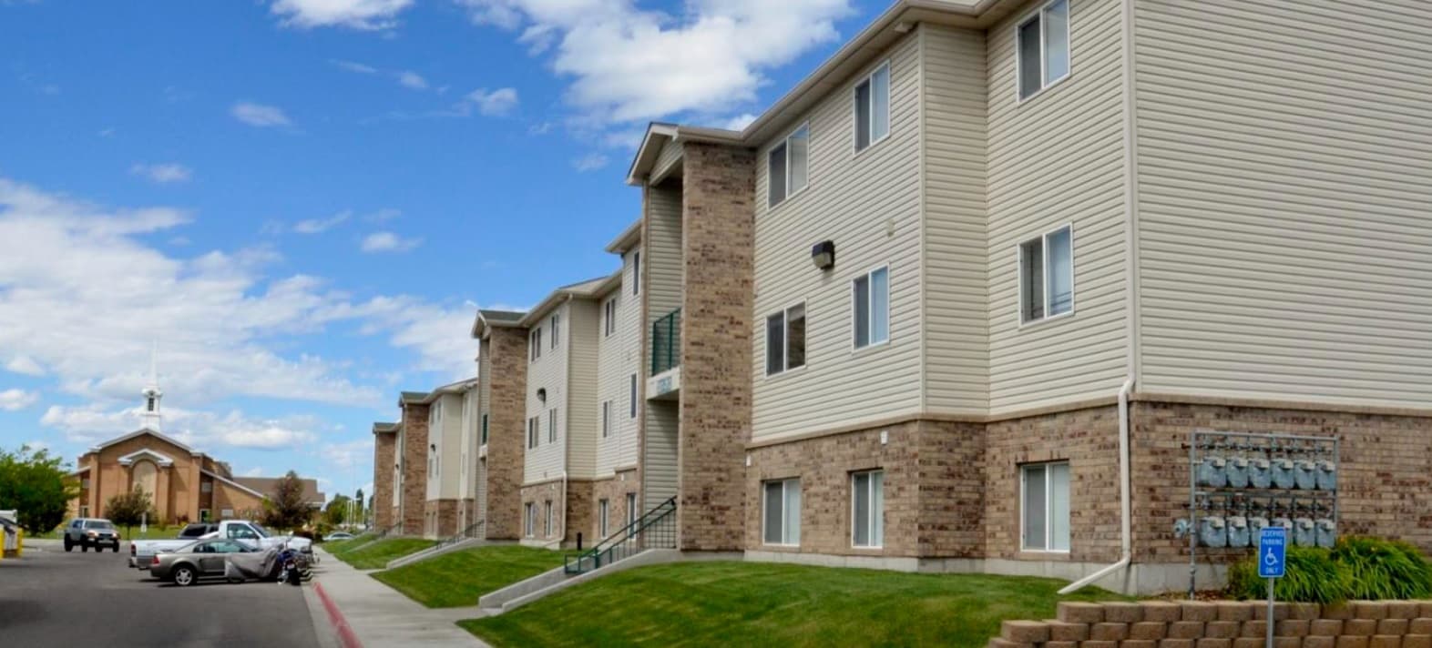 Cougar Court Apartments Rexburg is a three-story building with beige siding and brick exterior, green lawn, and a wheelchair-accessible parking space in the foreground; a church appears in the background under a partly cloudy sky.