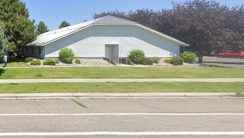 A white, single-story building with a gray metal roof sits behind a grassy lawn and sidewalk at Galaxy Rexburg, surrounded by bushes, small trees, and a parking lot. The sky is clear and sunny.