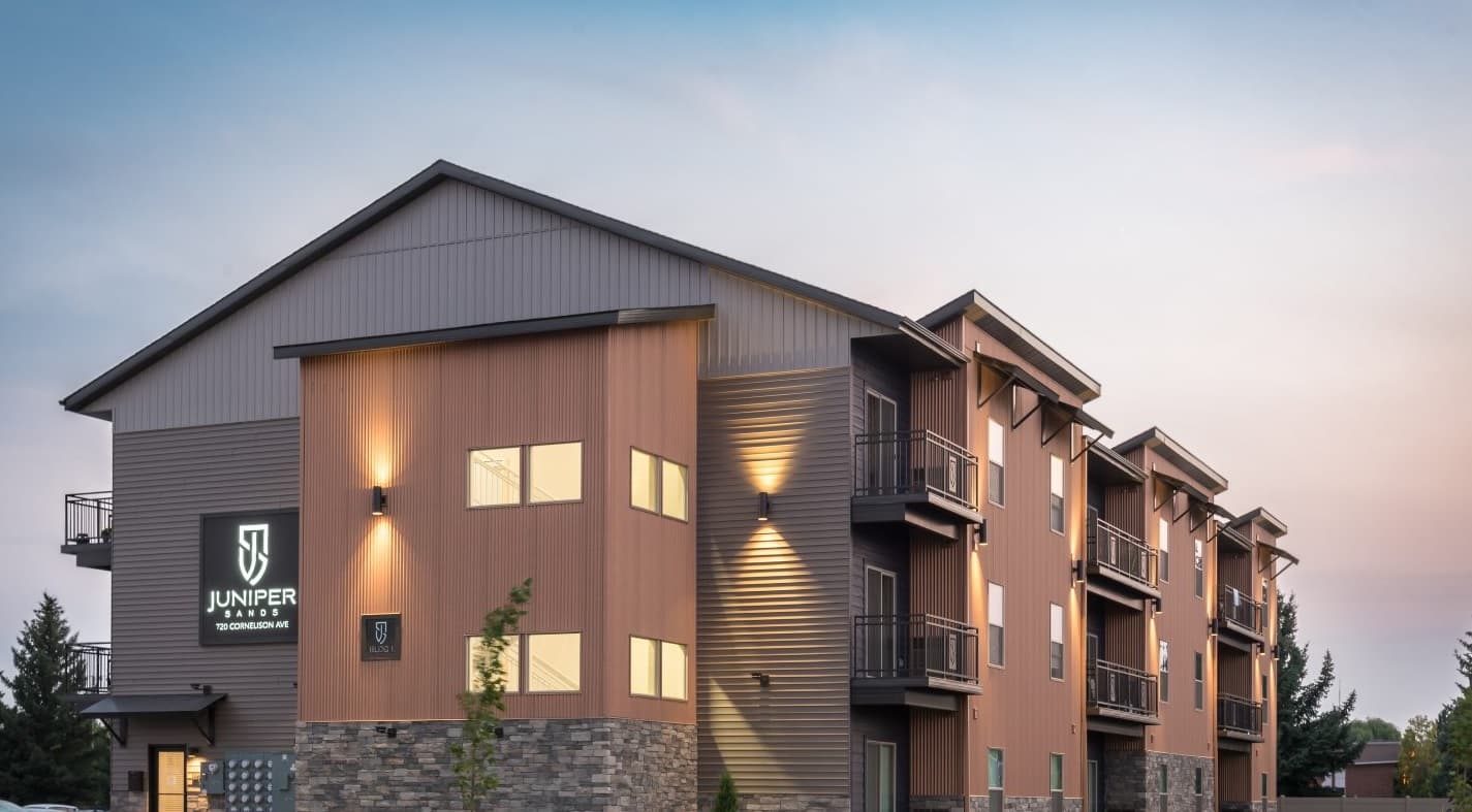 Modern three-story apartment building with wood and stone exterior, large windows, balconies, and a glowing “Juniper” sign on the corner, set against a pastel evening sky at Juniper Sands Rexburg.