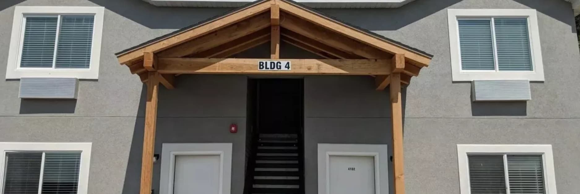 The entrance of Kenneth Square Apartments Rexburg, a gray building labeled BLDG 4, features a wooden awning, two white doors, and windows with closed blinds. A staircase leads up to the second floor.