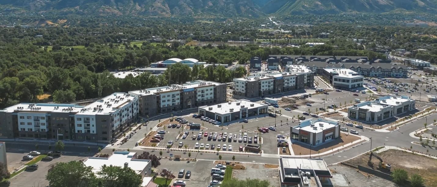 Aerial view of Lofts at River's Edge Provo, a modern apartment complex with multiple buildings, parking lots, and cars, surrounded by trees and mountains under a clear sky.