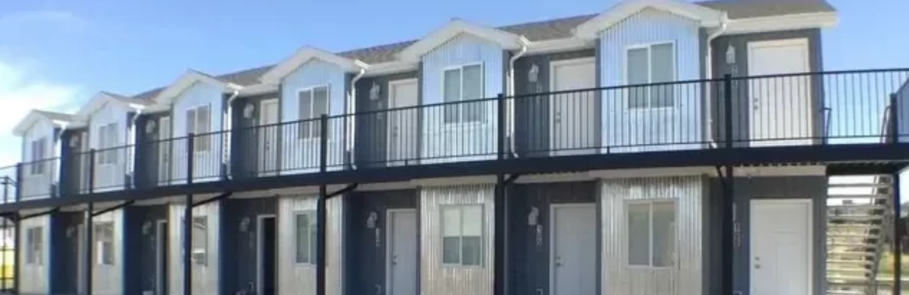 A two-story row of modern, small apartments at Teton Flats Rexburg features white doors, large windows, and a continuous black metal railing along the upper outdoor walkway under a clear sky.