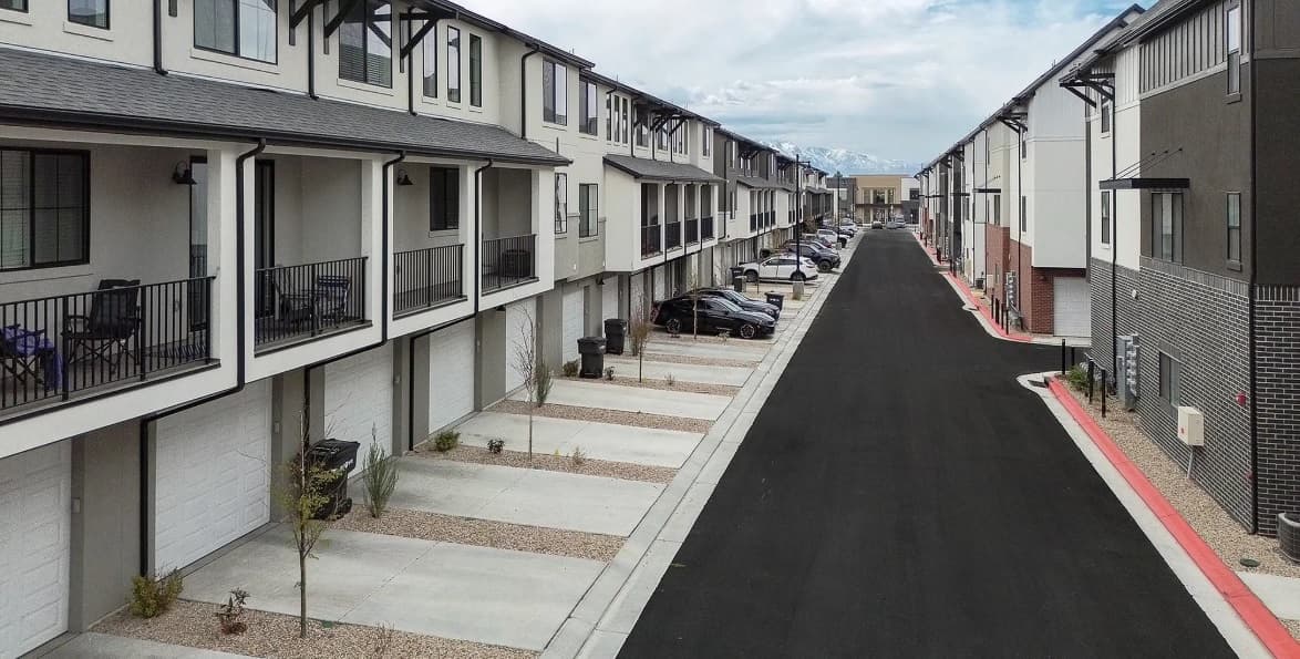 A row of modern Towns at River's Edge Provo townhouses with garages and balconies lines both sides of a clean, empty street on a clear day, with parked cars and mountains visible in the distance.