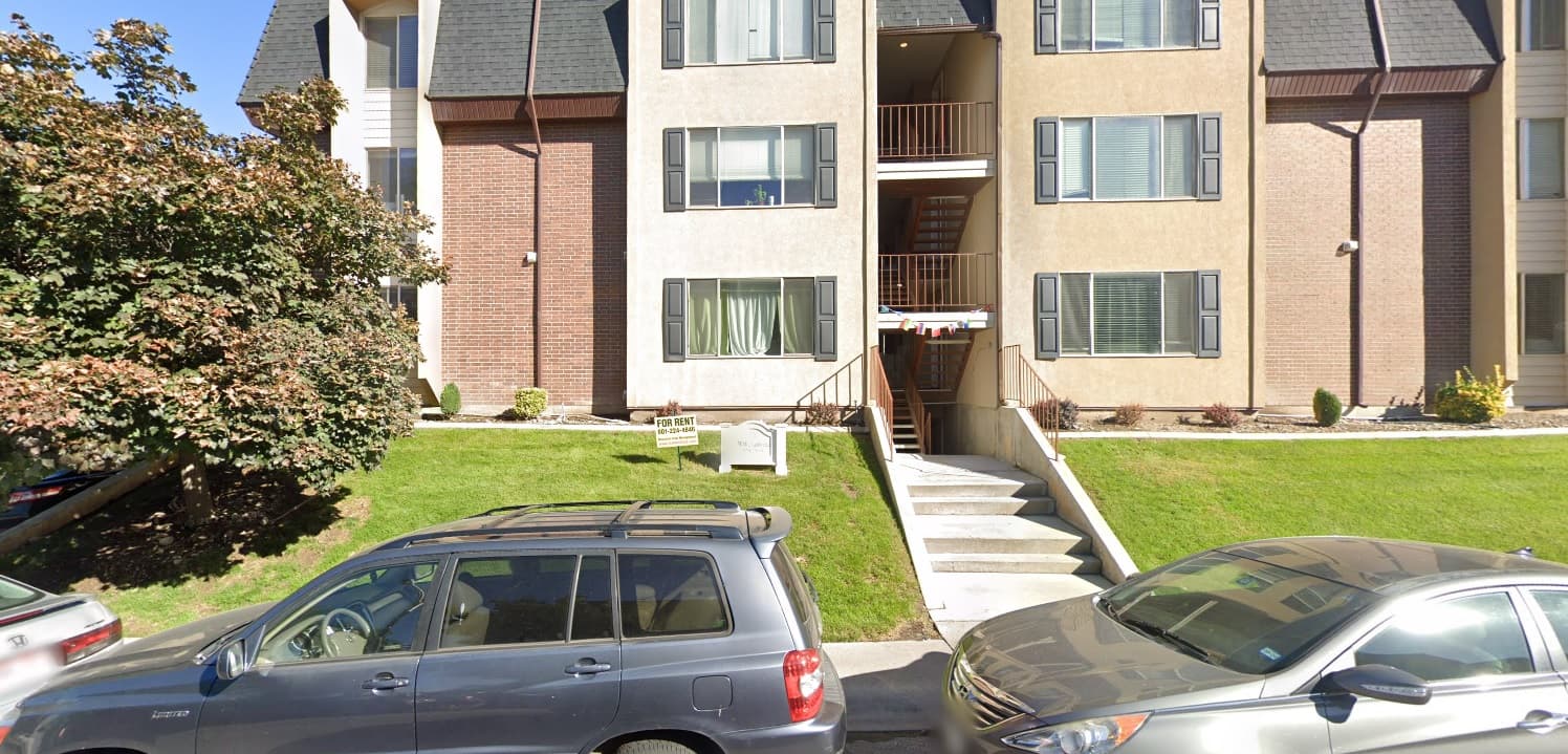 The Windsor Arms Provo is a tan and brick apartment building with three floors, a central stairway, green lawn, and parked cars in front on a sunny day.