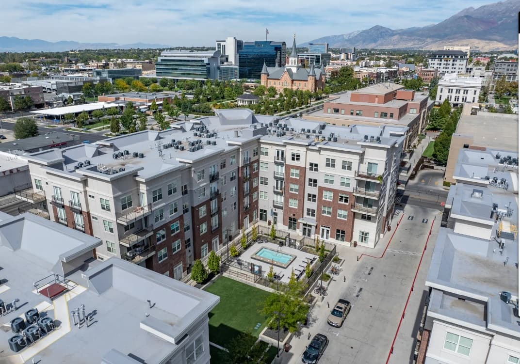 Aerial view of 200 City View Provo’s modern apartment buildings with a central courtyard and swimming pool, surrounded by city streets, office buildings, and mountains in the background under a clear sky.