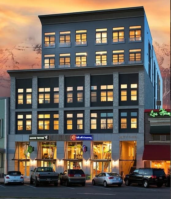 A modern, multi-story building with large windows illuminated from within, Sixty Three Center Provo features restaurants and shops at street level, several parked cars in front, and a scenic sunset with mountains in the background.