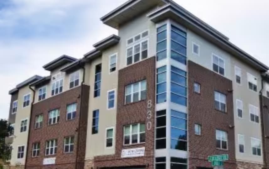 College 830 Apartments Fort Collins is a modern, four-story building with brick and beige siding, large windows, and a glass stairwell at the corner. The address “830” is visible along with street signs reading “Centerline Dr.”.