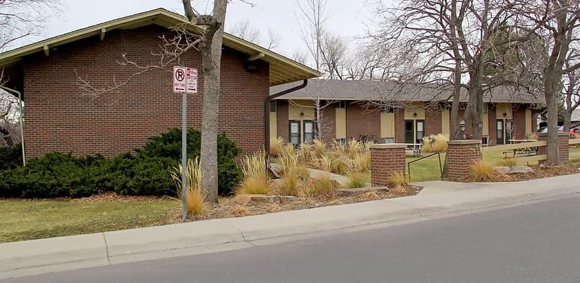 A one-story brick building with a sloped roof, surrounded by leafless trees, shrubs, ornamental grasses, and a sidewalk, sits near a no-parking sign—typical of the University Heights Apartments Boulder area.