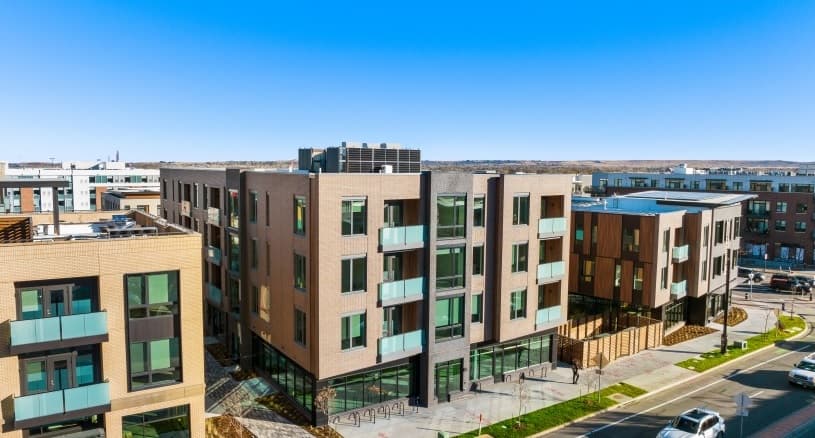 A modern, multi-story apartment building with glass balconies and large windows sits at a street corner in Boulder Commons Boulder, basking in the sun and surrounded by sidewalks and other contemporary buildings.