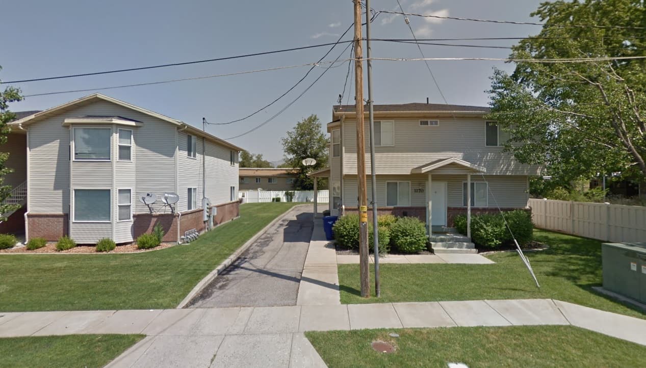 Two beige, two-story buildings at Brinker Apartments Ogden face a shared driveway lined with green lawns. Utility poles and wires run down the center, while trees and shrubs surround the apartments under a clear sky.