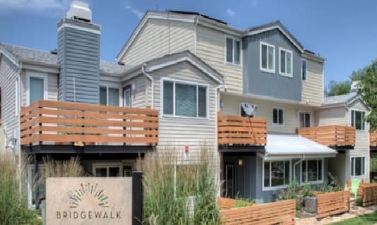 A modern multi-story townhouse complex, Bridgewalk Boulder features beige and gray siding, wooden balcony railings, and large windows. In the foreground, a sign reading Bridgewalk is surrounded by lush landscaping.