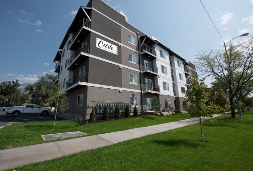 A modern four-story apartment building, The Carlo at Washington Ogden, features balconies, a gray and white exterior, and a "Condo" sign. Green grass, trees, and a sidewalk line the foreground beneath a blue sky.