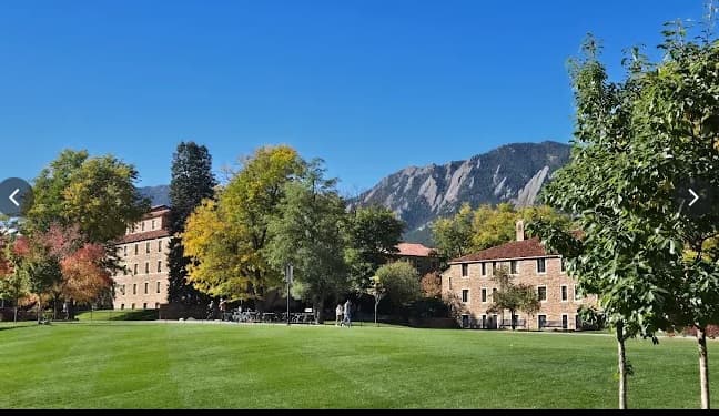 A large grassy lawn bordered by trees with autumn foliage stretches in front of tan brick buildings, including Crosman Hall. In the background, rocky mountains rise under a clear blue Boulder sky.