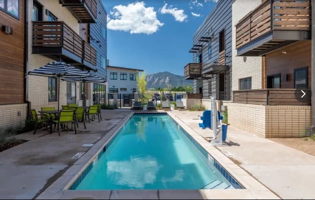 A narrow outdoor swimming pool at The Ellipse Boulder is surrounded by modern apartment buildings with balconies, patio tables, chairs, and umbrellas. Mountains and a blue sky with clouds are visible in the background.