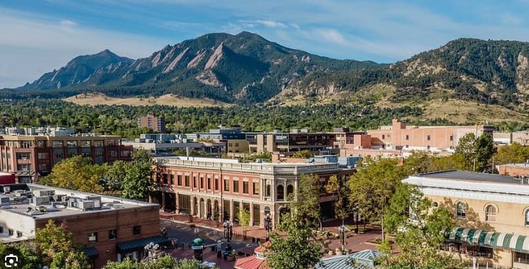 A view of a small city with brick buildings and tree-lined streets, set against green foothills and tall, rugged mountains under a clear blue sky—discover the charm Housing Helpers Boulder brings to this picturesque setting.