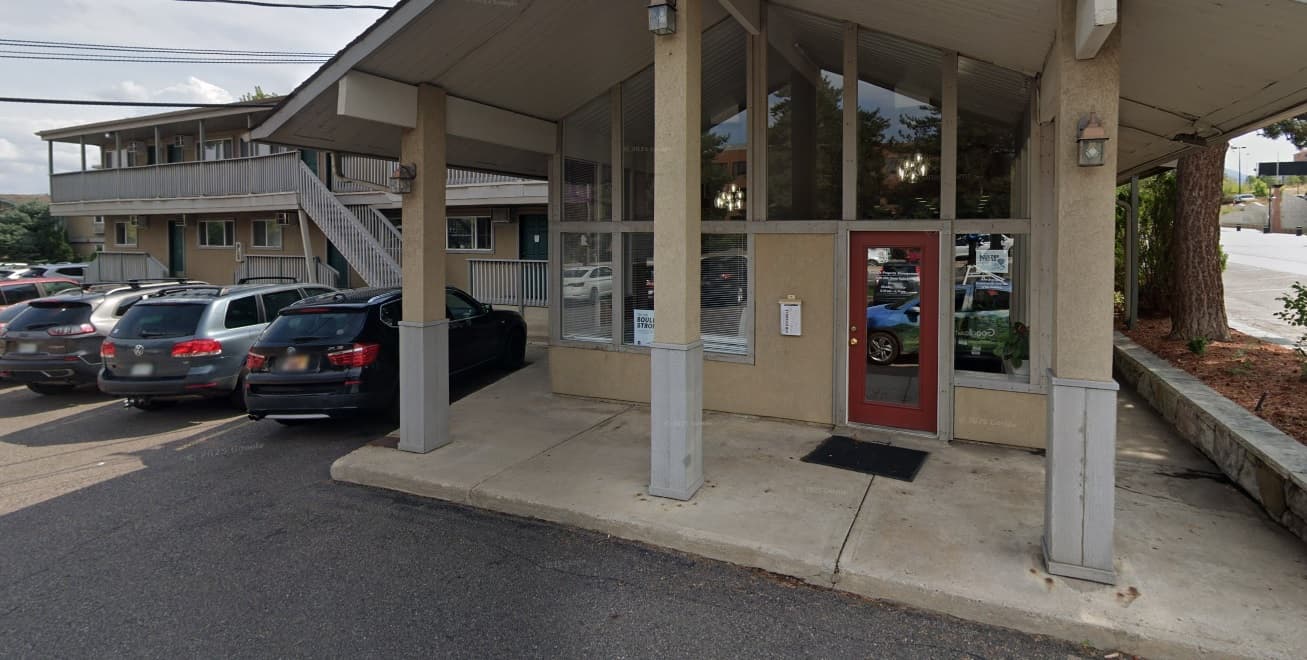 A small office entrance with a red door and large windows under an overhanging roof sits beside Jay’s Apartment Boulder, next to a parking lot with several parked cars. An outdoor staircase leads to an upper level in the background.