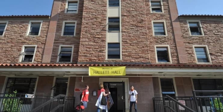 Three people stand outside the entrance of a stone dormitory building with a yellow sign reading Welcome to Hallett Hall above the doors, near Libby Hall on CU Boulder's campus. The building has multiple windows and a red-tiled roof.