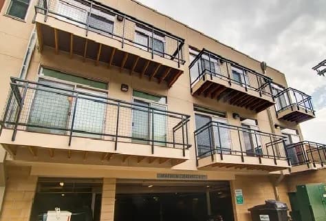 Lofts On College Boulder is a modern apartment building with several small balconies featuring metal railings, large windows, and a parking garage entrance below, all set under a cloudy sky.