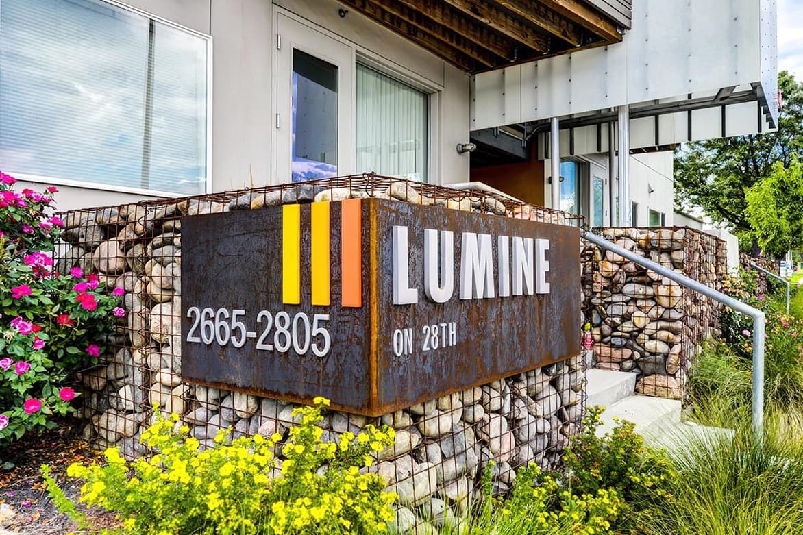 A modern building entrance with a rust-colored sign reading “LUMINE ON 28TH” and “2665-2805” in white text, marking Lumine Apartments Boulder, is framed by gabion rock walls and blooming flowers beneath a wooden awning.