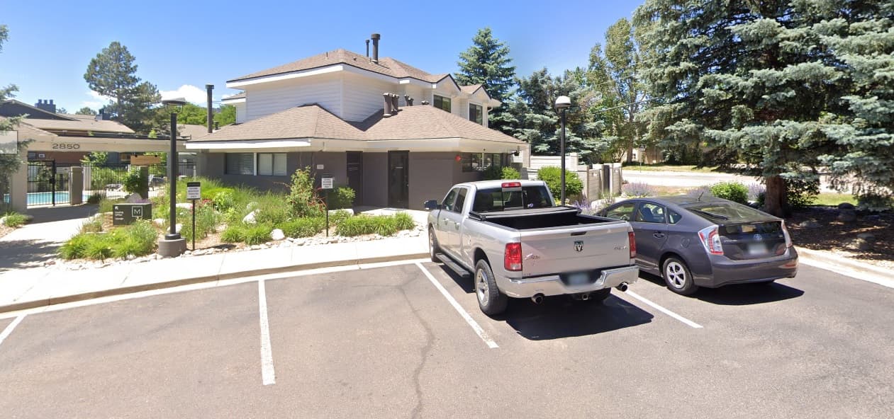 The Magwood Apartments Boulder is a white two-story building with a covered entrance, surrounded by trees and greenery. A silver pickup truck and a gray sedan are parked in front of this inviting residence on a sunny day.