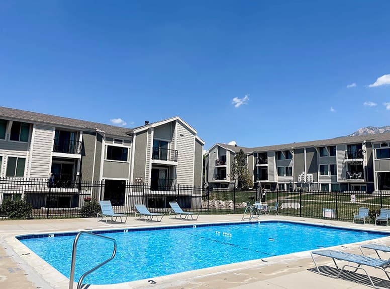 A bright outdoor pool at The Rainier Ogden, surrounded by lounge chairs, sits in front of two-story apartment buildings under a clear blue sky. A black fence encloses the inviting pool area.