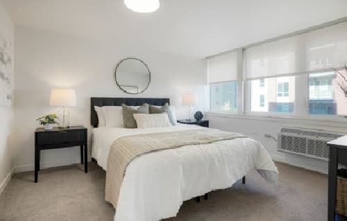 A modern bedroom at Red Oak Park Boulder features a neatly made bed with white bedding, a beige throw blanket, two black nightstands with lamps, a round mirror above the bed, large windows, and neutral decor.