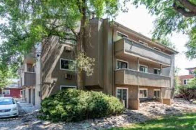 A three-story Spruce Street Apartments Boulder building with beige walls and large balconies, surrounded by trees and bushes, with a white car parked nearby and sunlight filtering through the leaves.