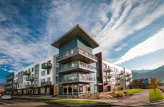 Modern four-story apartment building with large balconies and glass windows stands at Startup Crossing Provo, on a street corner under a blue sky with wispy clouds. Trees and a small lawn add to the inviting foreground.
