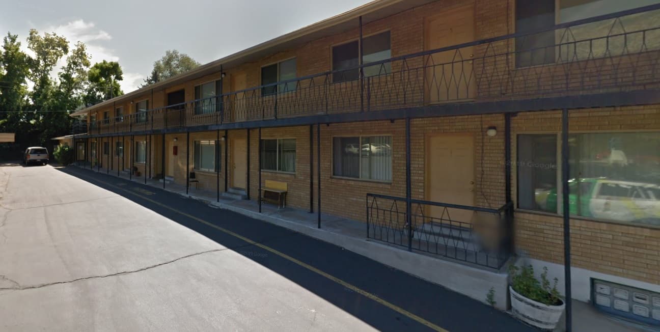 A two-story brick motel, similar to Tamlyn Apartments Ogden, features exterior room doors with metal railings, large windows with blinds, and a potted plant by one entrance. Each door faces a paved driveway, with trees visible in the background.