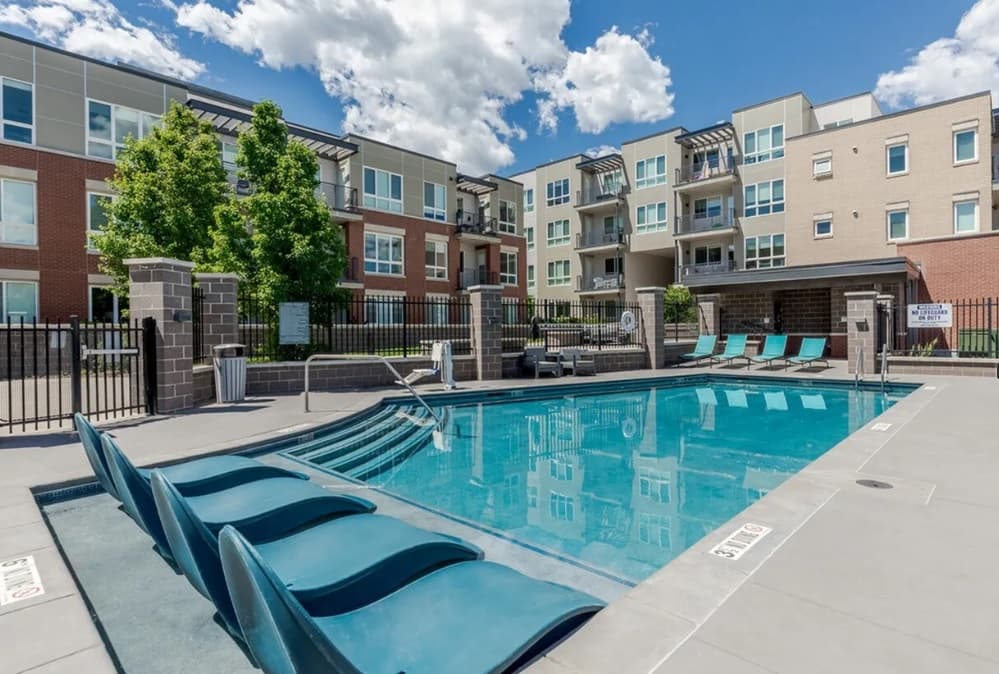 Modern Union 9Seventy Boulder apartment complex with a fenced outdoor pool, several lounge chairs partially in the water, and multi-story buildings with balconies and large windows under a blue sky with clouds.