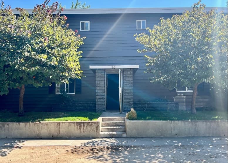 A two-story dark gray building with white trim, stone columns at the entrance, and two trees flanking the entryway, 310 Thatcher Apartments Boise offers inviting concrete steps leading to the front door as sunlight creates a glare on the right side.