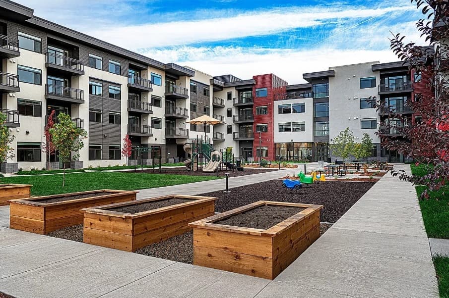 Adare Boise is a modern apartment complex with balconies overlooking a courtyard featuring raised garden beds, a playground with slides and spring riders, trees, and paved walkways under a partly cloudy sky.