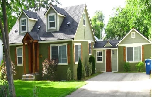The Agate House Boise: A green house with white trim and brown shutters, featuring a front porch, dormer windows, a driveway, and a blue recycling bin on the right side, surrounded by lush trees and grass.