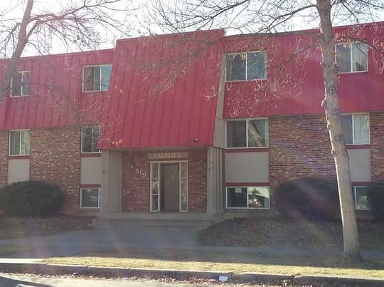 A brick apartment building with a red roof and trim, labeled CAVALIER above the entrance. The glass doors mark the entry to Cavalier Apartments Fort Collins, with leafless trees and shrubs in front.