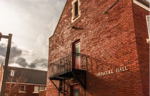 A brick building with a small balcony and a door on the second floor. The sign on the wall reads Driscoll Hall Boise. The sky is cloudy, and another brick building is visible in the background.