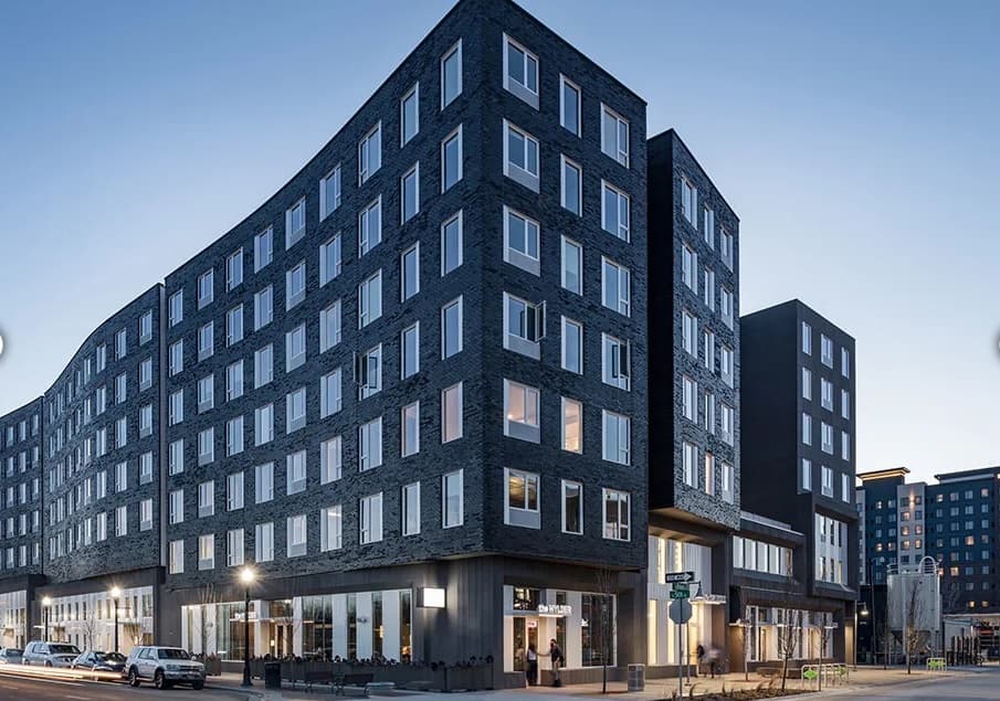 The Fowler Apartments Boise is a modern, multi-story building with a dark brick exterior and large windows, photographed at dusk as cars line the street and lights glow warmly from the bustling ground-floor shops.