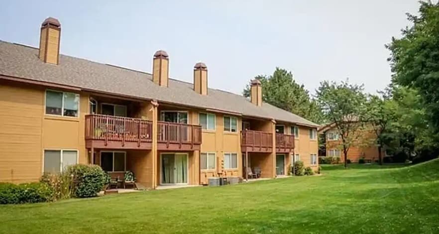 Two-story brown apartment building with balconies and patios at Gekeler Farms Boise, surrounded by green grass and trees on a clear day.