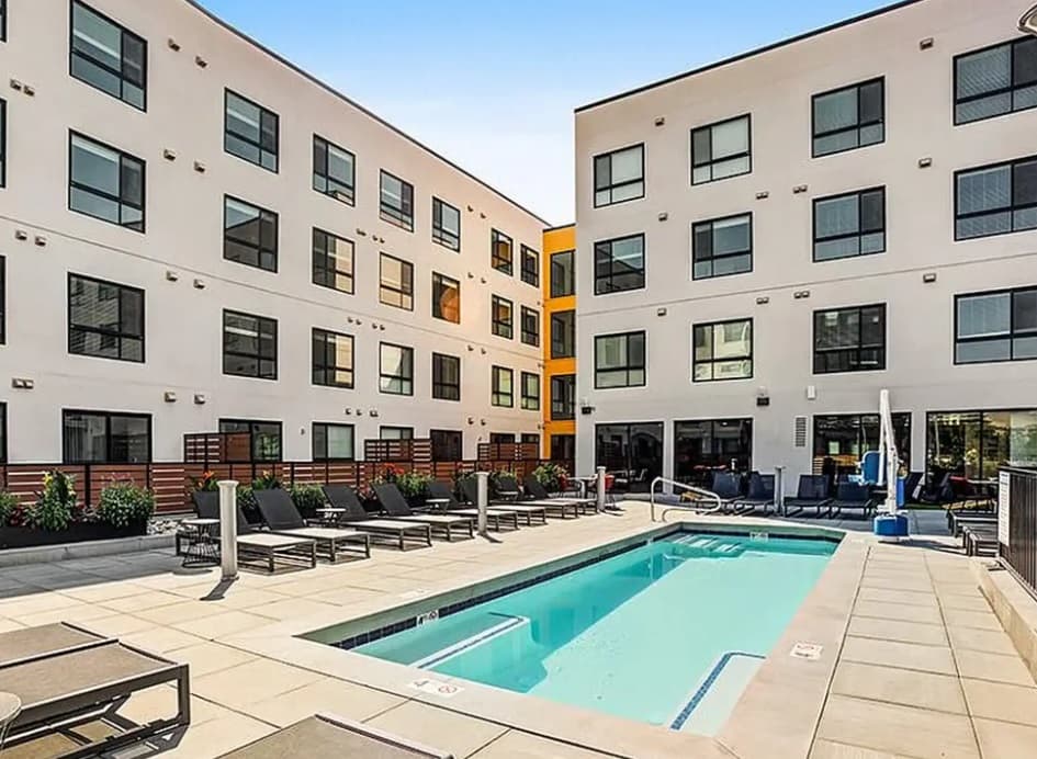 A modern courtyard at Jules on 3rd Boise features a rectangular swimming pool, surrounded by lounge chairs, potted plants, and tall white buildings with many windows under a clear sky.