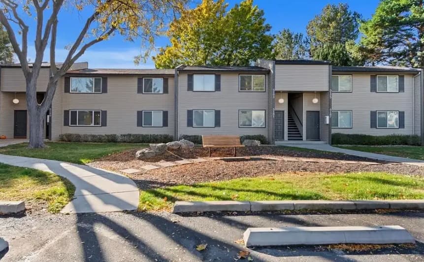 A two-story apartment building with gray siding at Juniper West Boise, surrounded by green grass, trees, and a mulch area with a bench; parking spaces and a sidewalk are in the foreground under a clear blue sky.