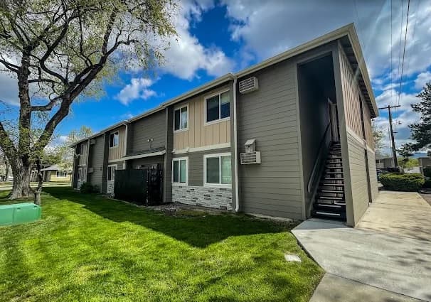 Jefferson West Boise two-story apartment building with beige and brown siding, stone accents, and outdoor staircase. A green lawn and trees surround the building under a partly cloudy sky.