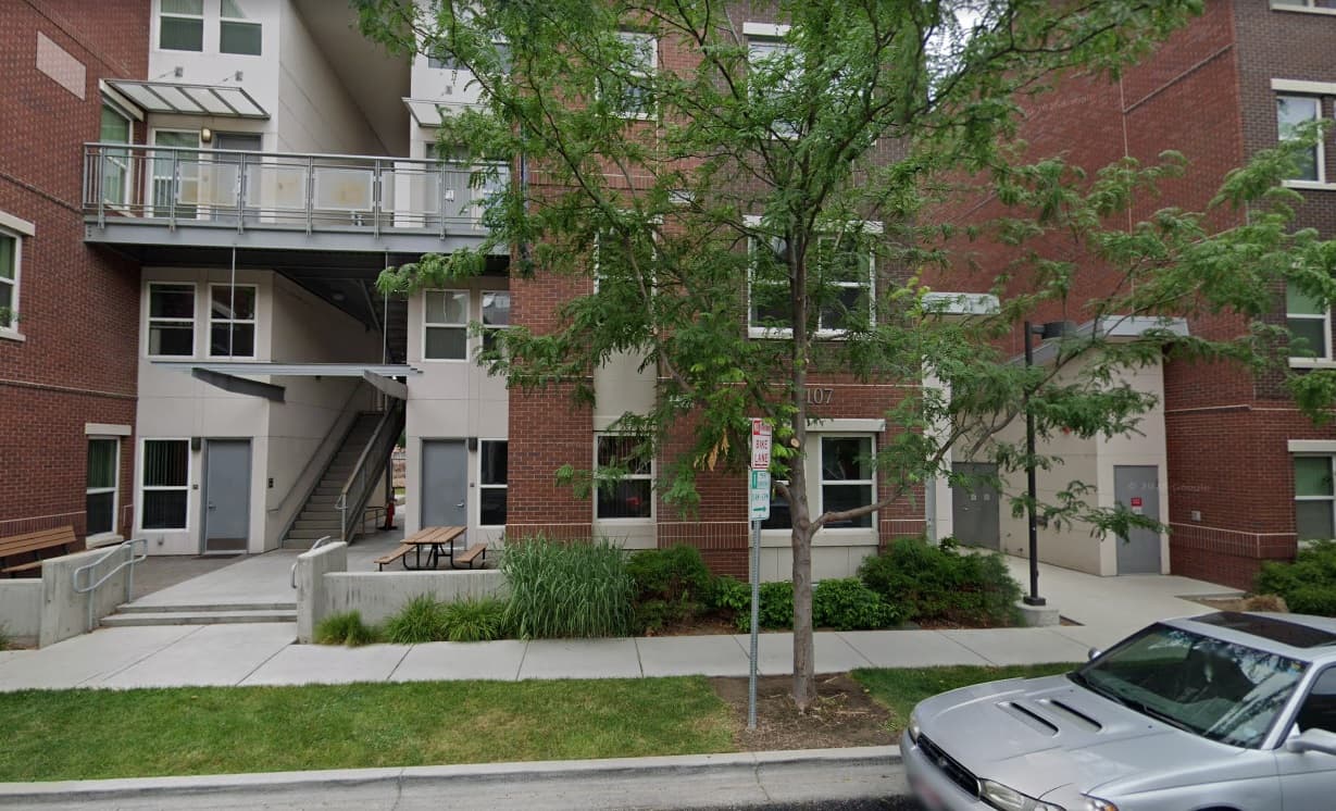 Lincoln Townhomes Boise features a modern apartment building with red brick and beige siding, balconies, and an external staircase. Trees, greenery, a sidewalk, and a parked silver car enhance the inviting atmosphere out front.