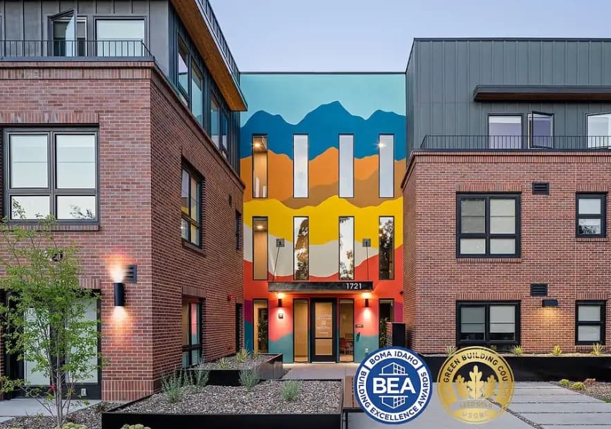 A modern apartment building, The Martha Apartments Boise features a colorful mural of mountains at the front entrance, surrounded by large windows and brick walls. Two award badges are displayed in the lower right corner.