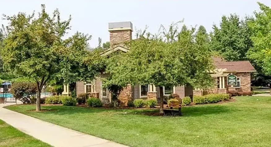A small brick and wood building with a chimney sits amid green grass, trees, and shrubs in Oak Park Village Boise. A pathway and bench are in the foreground, with a swimming pool partially visible to the left.